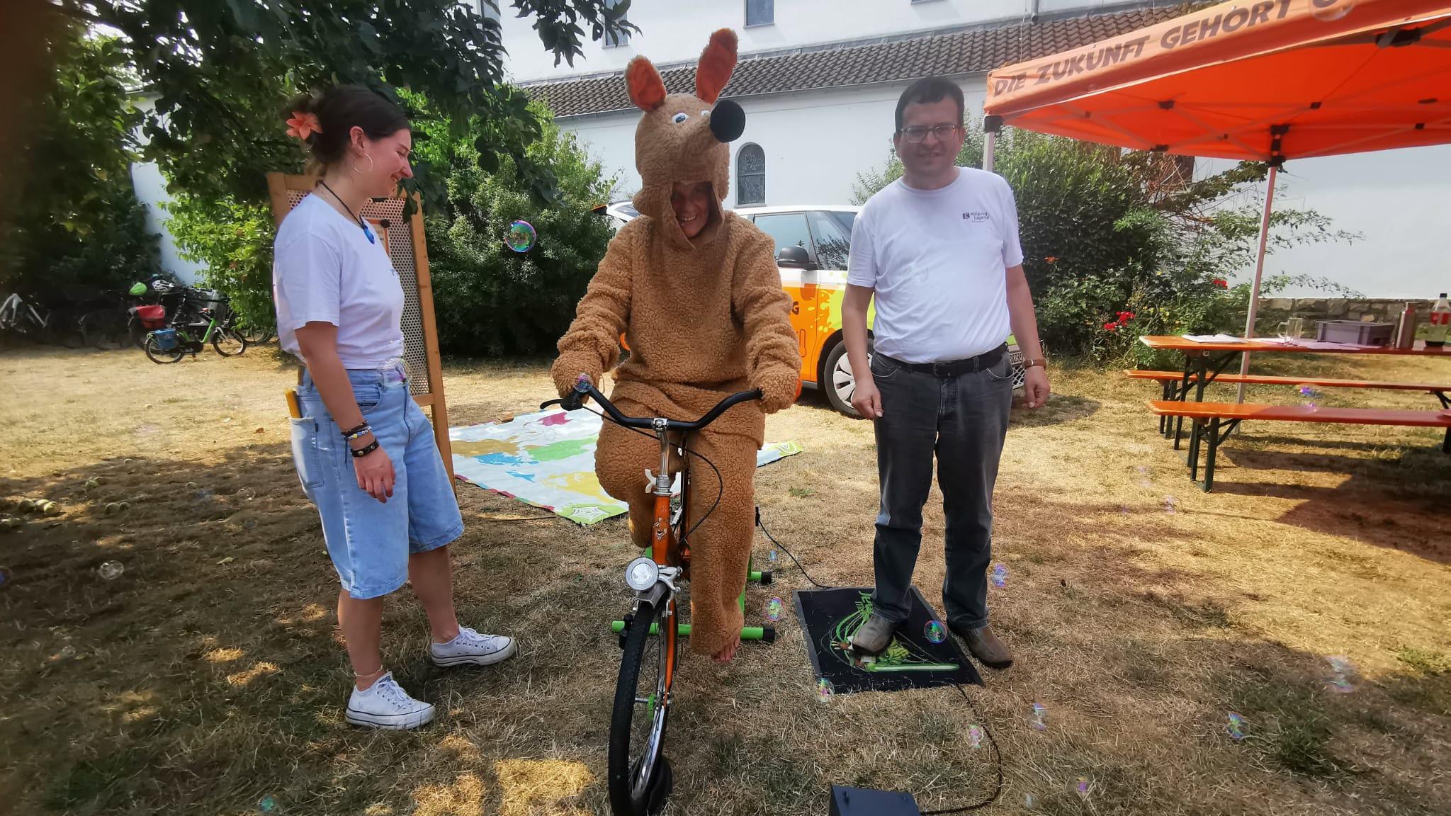 Schnuffi das Maskotchen auf dem Fahrrad, dazu zwei ehrenamtliche Mitarbeiter